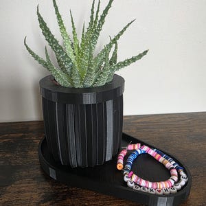 May include: A black, ribbed planter pot holding a green succulent plant, sitting on a matching black tray. A beaded bracelet with the word "PUERTO RICO" is on the tray. The items are on a dark wood surface.