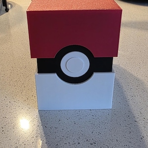 May include: A decorative box designed to resemble a Poké Ball. The top is red with glitter, the middle is black with a white circle, and the bottom is white. The box is sitting on a speckled countertop.