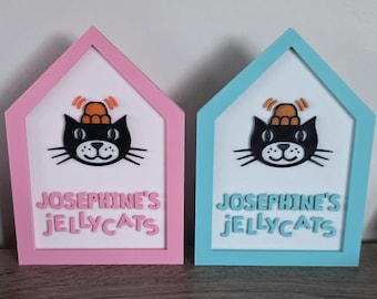 May include: Two house-shaped signs with white backgrounds. The left sign has a pink frame and pink text reading "JOSEPHINE'S JELLYCATS". The right sign has a light blue frame and blue text reading "JOSEPHINE'S JELLYCATS". Both feature a black cat icon wearing an orange hat.