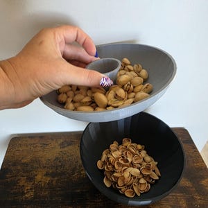 Puede incluir: Un cuenco gris impreso en 3D con un compartimento central, lleno de pistachos, se sostiene sobre un cuenco negro lleno de cáscaras de pistacho. Los cuencos están sobre una superficie de madera. El diseño es moderno y funcional.