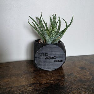 May include: A small, black, cylindrical planter with a succulent plant. The planter has a circular design with the words "TALK dirt TOME" printed on it. The succulent has green, spiky leaves with white spots.