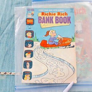 May include: A vintage Richie Rich Bank Book comic book, issue number 9, priced at 20 cents. The cover features a cartoon character riding a red snowmobile on a path marked with dollar signs, with small character portraits along the left side.