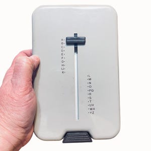 May include: A rectangular light gray plastic device featuring a vertical sliding black tab in a central track. The surface displays two columns of letters from A to Z, with dots next to each letter. A dark gray textured tab is located at the bottom edge.