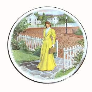 Puede incluir: Un plato decorativo redondo con una ilustración colorida de una persona con un abrigo largo amarillo y un sombrero marrón, de pie en un camino de piedra cerca de una valla blanca, con casas y una farola visibles en el fondo.