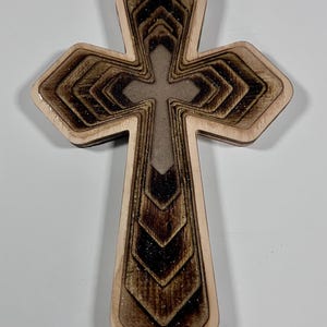 Può includere: Croce in legno con un design a strati. La croce presenta un bordo marrone chiaro con un interno strutturato marrone più scuro. La croce centrale è marrone chiaro. Il design ha una finitura glitterata. Arredamento religioso.