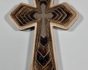 May include: Wooden cross with a layered design. The cross features a light brown border with a darker brown, textured interior. The center cross is a lighter brown. The design has a glittery finish. Religious decor.