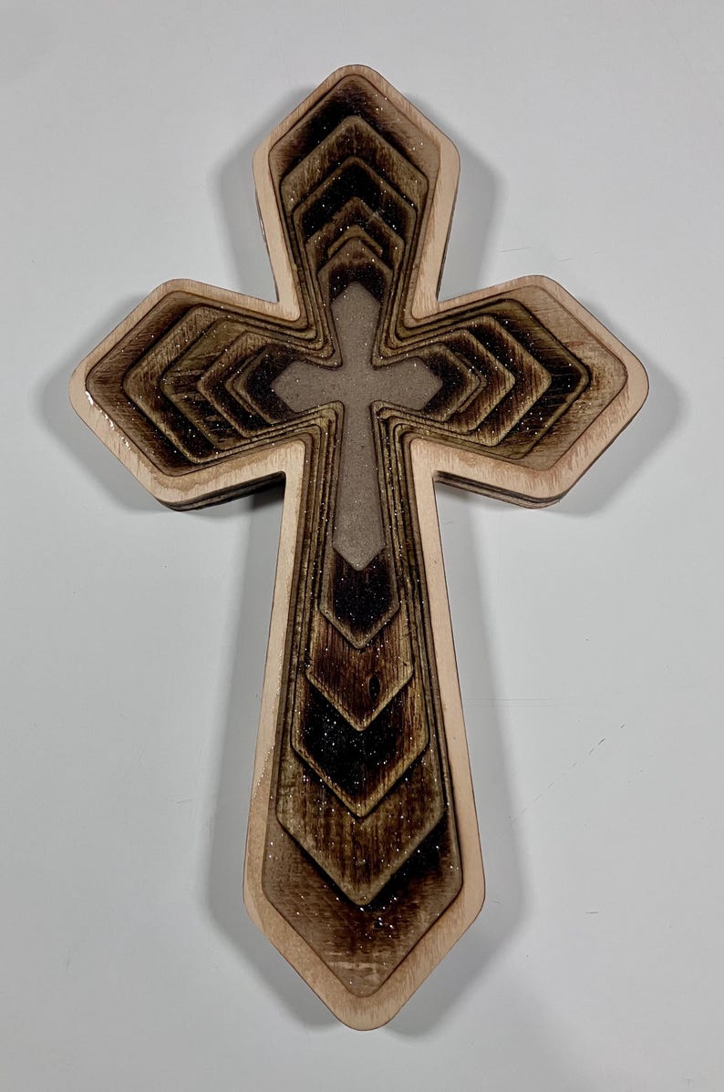 May include: Wooden cross with a layered design. The cross features a light brown border with a darker brown, textured interior. The center cross is a lighter brown. The design has a glittery finish. Religious decor.