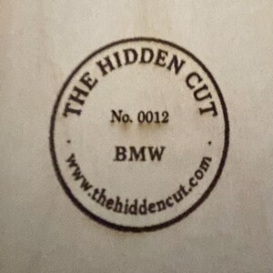 Puede incluir: Sello circular con el texto "THE HIDDEN CUT" en la parte superior y "BMW" en la inferior. El n&uacute;mero "0012" est&aacute; en el centro. La direcci&oacute;n del sitio web "www.thehidencut.com" est&aacute; en la parte inferior del sello.