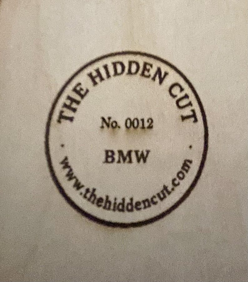 Puede incluir: Sello circular con el texto "THE HIDDEN CUT" en la parte superior y "BMW" en la inferior. El n&uacute;mero "0012" est&aacute; en el centro. La direcci&oacute;n del sitio web "www.thehidencut.com" est&aacute; en la parte inferior del sello.