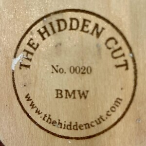 May include: Circular wooden emblem with the text "THE HIDDEN CUT" in a circular pattern. The text "No. 0020" and "BMW" are also visible. The website address www.thehiddencut.com is printed at the bottom of the emblem.