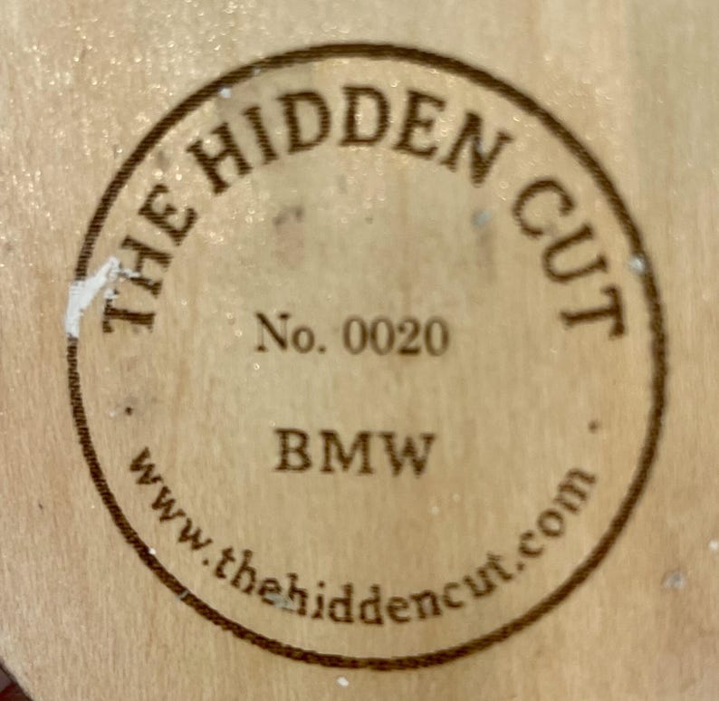 May include: Circular wooden emblem with the text "THE HIDDEN CUT" in a circular pattern. The text "No. 0020" and "BMW" are also visible. The website address www.thehiddencut.com is printed at the bottom of the emblem.