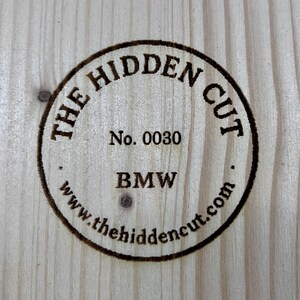 May include: Circular wooden emblem with the text "THE HIDDEN CUT" and "BMW" in a dark brown color. The emblem also includes the number "0030" and the website address "www.thehiddencut.com". The emblem is set against a light wood grain background.
