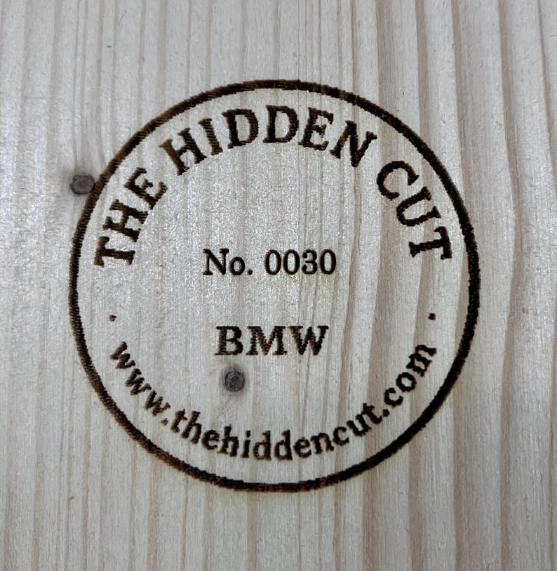 May include: Circular wooden emblem with the text "THE HIDDEN CUT" and "BMW" in a dark brown color. The emblem also includes the number "0030" and the website address "www.thehiddencut.com". The emblem is set against a light wood grain background.