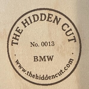 May include: A circular wooden emblem with the text "THE HIDDEN CUT" in a ring around the edge. Inside the circle are the words "No. 0013" and "BMW", with the website address www.thehiddencut.com at the bottom.