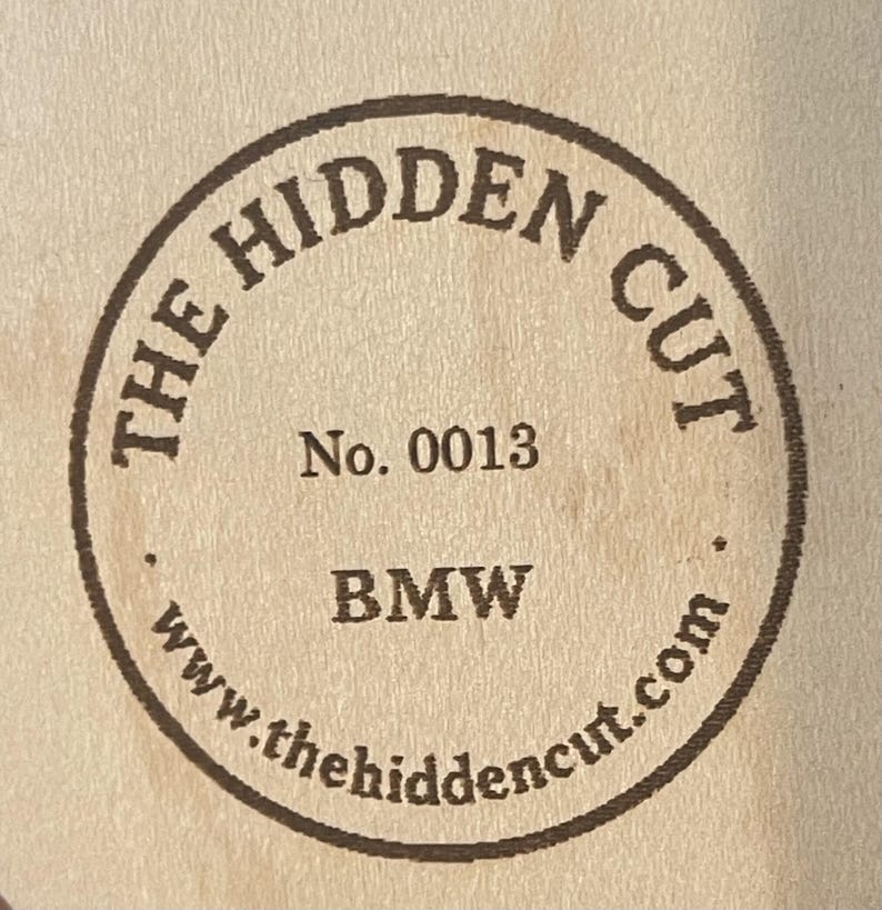 May include: A circular wooden emblem with the text "THE HIDDEN CUT" in a ring around the edge. Inside the circle are the words "No. 0013" and "BMW", with the website address www.thehiddencut.com at the bottom.
