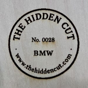 May include: Circular wooden emblem with the text "THE HIDDEN CUT" in a curved font. The number "0028" is centered below, followed by "BMW" and the website address "www.thehiddencut.com" in a circular pattern.