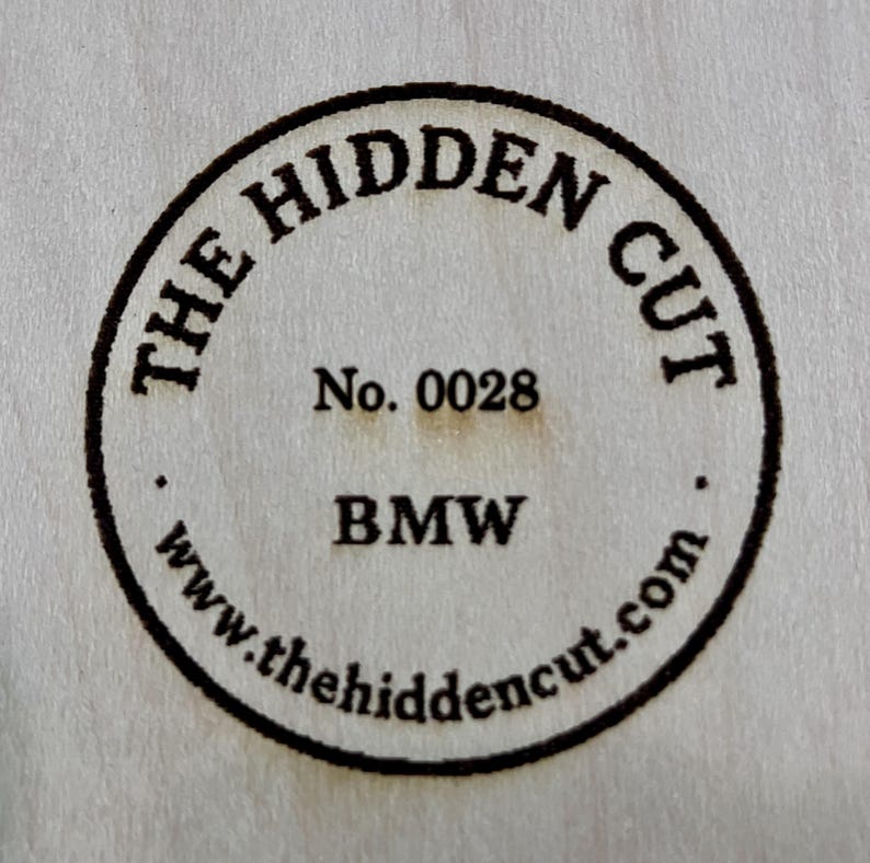 May include: Circular wooden emblem with the text "THE HIDDEN CUT" in a curved font. The number "0028" is centered below, followed by "BMW" and the website address "www.thehiddencut.com" in a circular pattern.