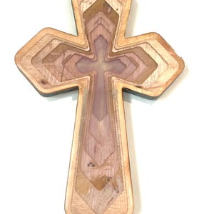 Può includere: Una croce in legno a strati con un esterno marrone chiaro e un interno multicolore. La croce presenta un disegno a forma di croce al centro e gli strati creano un effetto dimensionale. La venatura del legno è visibile.