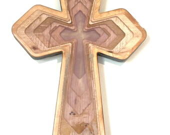 May include: A layered wooden cross with a light brown exterior and a multi-toned interior. The cross features a cross-shaped design within its center, and the layers create a dimensional effect. The wood grain is visible throughout the design.