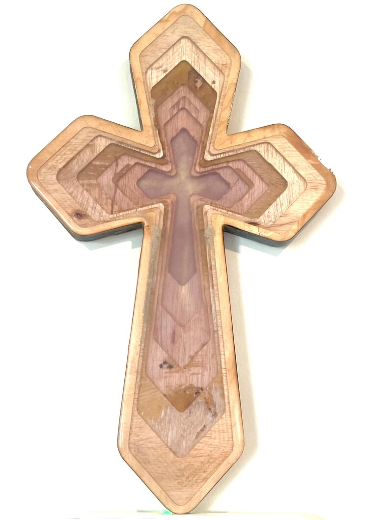 May include: A layered wooden cross with a light brown exterior and a multi-toned interior. The cross features a cross-shaped design within its center, and the layers create a dimensional effect. The wood grain is visible throughout the design.