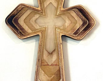 May include: Wooden cross with a layered design. The cross features a light-colored center and a darker, stained wood border. The design incorporates geometric shapes and a cross-shaped cutout in the center.
