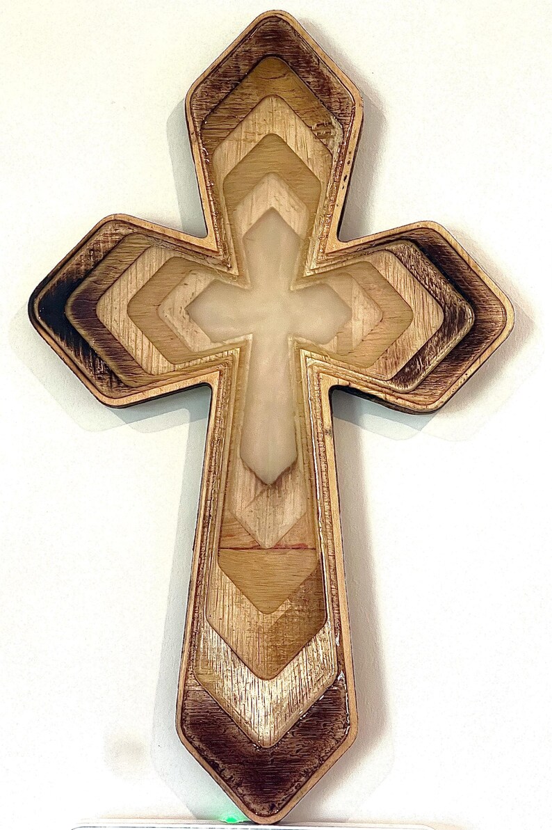 Puede incluir: Cruz de madera con un dise&ntilde;o en capas. La cruz presenta un centro de color claro y un borde de madera te&ntilde;ida m&aacute;s oscuro. El dise&ntilde;o incorpora formas geom&eacute;tricas y un recorte en forma de cruz en el centro.