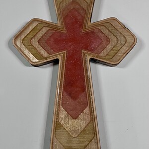 May include: Wooden cross with a layered design. The cross features a red center with glitter and is framed by alternating layers of light and dark wood. The edges are smooth and the overall shape is symmetrical.