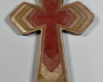 May include: Wooden cross with a layered design. The cross features a red center with glitter and is framed by alternating layers of light and dark wood. The edges are smooth and the overall shape is symmetrical.
