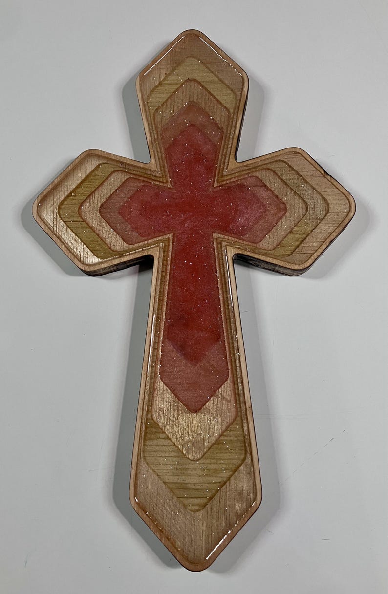 May include: Wooden cross with a layered design. The cross features a red center with glitter and is framed by alternating layers of light and dark wood. The edges are smooth and the overall shape is symmetrical.
