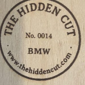 May include: Circular wooden emblem with the text "THE HIDDEN CUT" in a circular pattern. The text "No. 0014" and "BMW" are centered. The website address "www.thehiddencut.com" is also included in the design.
