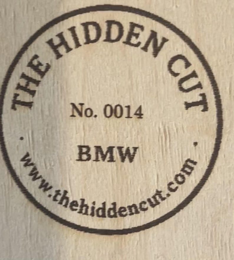 May include: Circular wooden emblem with the text "THE HIDDEN CUT" in a circular pattern. The text "No. 0014" and "BMW" are centered. The website address "www.thehiddencut.com" is also included in the design.
