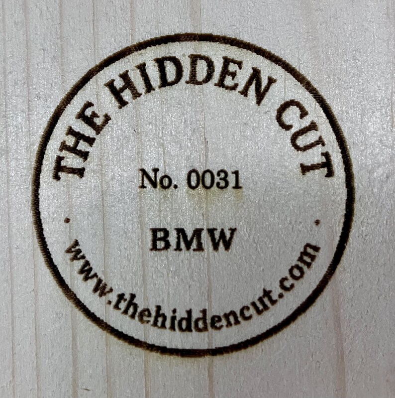 Pu&ograve; includere: Emblema circolare in legno con la scritta "THE HIDDEN CUT" sul perimetro. Il centro include "No. 0031" e "BMW" sotto. &Egrave; visibile anche l'indirizzo del sito web "www.thehiddencut.com".