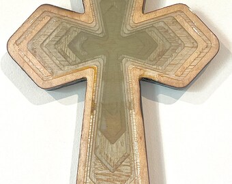 May include: Wooden cross with a layered design. The cross features a light brown exterior with a series of interior layers, creating a three-dimensional effect. The center of the cross is a lighter, translucent color. The cross is a religious symbol.
