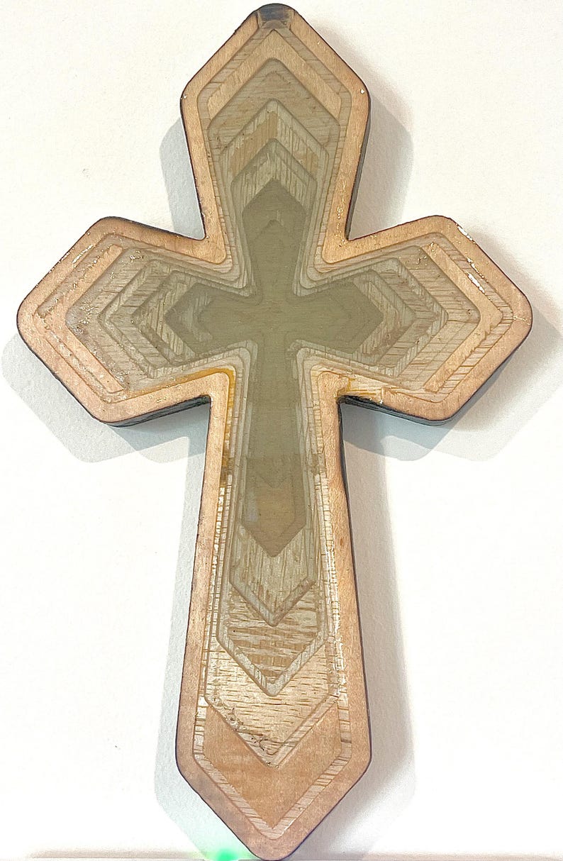 May include: Wooden cross with a layered design. The cross features a light brown exterior with a series of interior layers, creating a three-dimensional effect. The center of the cross is a lighter, translucent color. The cross is a religious symbol.
