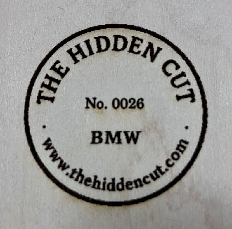May include: Circular emblem with the text "THE HIDDEN CUT" in a ring around the edge. The text "No. 0026" and "BMW" are in the center. The website address "www.thehiddencut.com" is at the bottom of the circle.