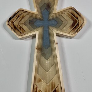Pu&ograve; includere: Croce in legno con design a strati. La croce presenta un centro azzurro chiaro ed &egrave; circondata da strati di legno in varie tonalit&agrave; di marrone e beige. I bordi del legno hanno un aspetto bruciato.
