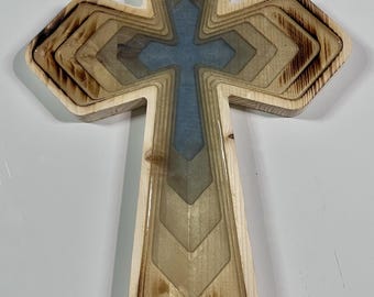 May include: Wooden cross with a layered design. The cross features a light blue center and is surrounded by layers of wood in varying shades of brown and tan. The edges of the wood have a burnt appearance.