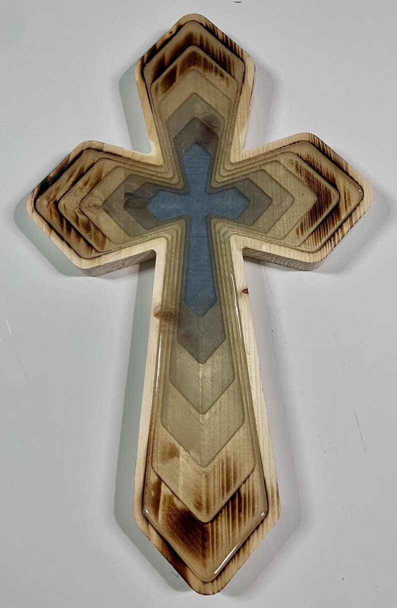 Pu&ograve; includere: Croce in legno con design a strati. La croce presenta un centro azzurro chiaro ed &egrave; circondata da strati di legno in varie tonalit&agrave; di marrone e beige. I bordi del legno hanno un aspetto bruciato.