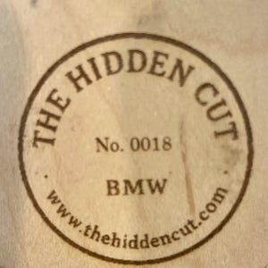 May include: A circular wooden emblem with the text "THE HIDDEN CUT" in a brown, circular border. The text "No. 0018" and "BMW" are centered within the circle. The website address "www.thehiddencut.com" is at the bottom.