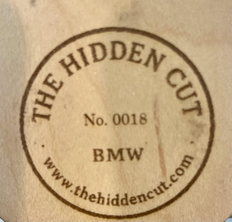 May include: A circular wooden emblem with the text "THE HIDDEN CUT" in a brown, circular border. The text "No. 0018" and "BMW" are centered within the circle. The website address "www.thehiddencut.com" is at the bottom.