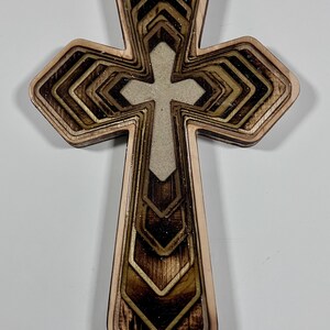 May include: A layered wooden cross with a light-colored center cross. The outer layers are a light brown, with darker brown and gold accents. The cross has a geometric design and is likely for religious decor.