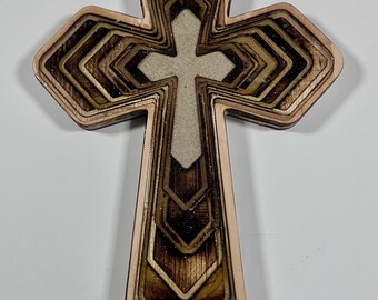 May include: A layered wooden cross with a light-colored center cross. The outer layers are a light brown, with darker brown and gold accents. The cross has a geometric design and is likely for religious decor.