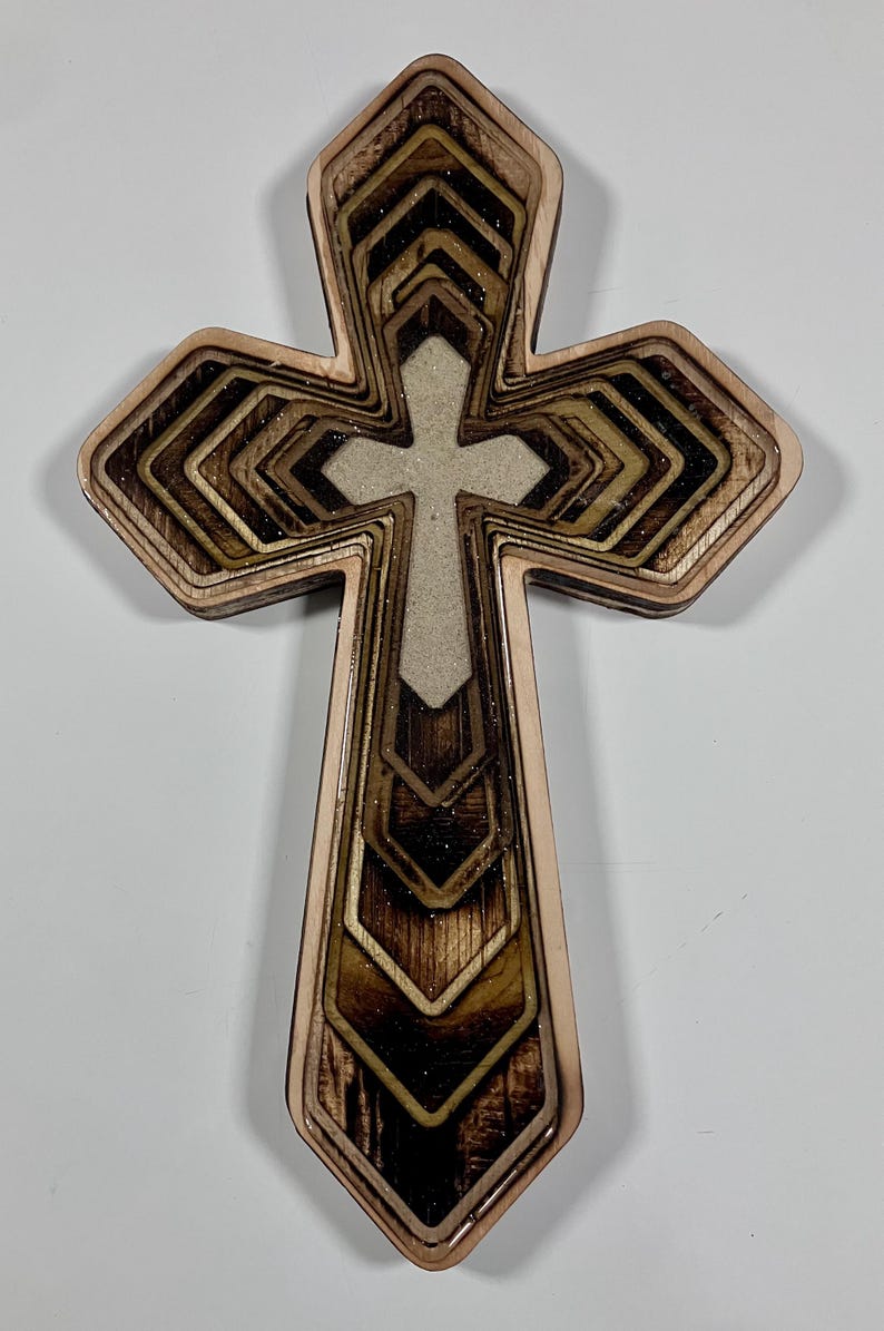 May include: A layered wooden cross with a light-colored center cross. The outer layers are a light brown, with darker brown and gold accents. The cross has a geometric design and is likely for religious decor.