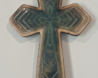 May include: Wooden cross with a layered design. The cross features a light brown border and a central design with shades of green and brown. The cross has a detailed, geometric pattern and is likely a decorative item.