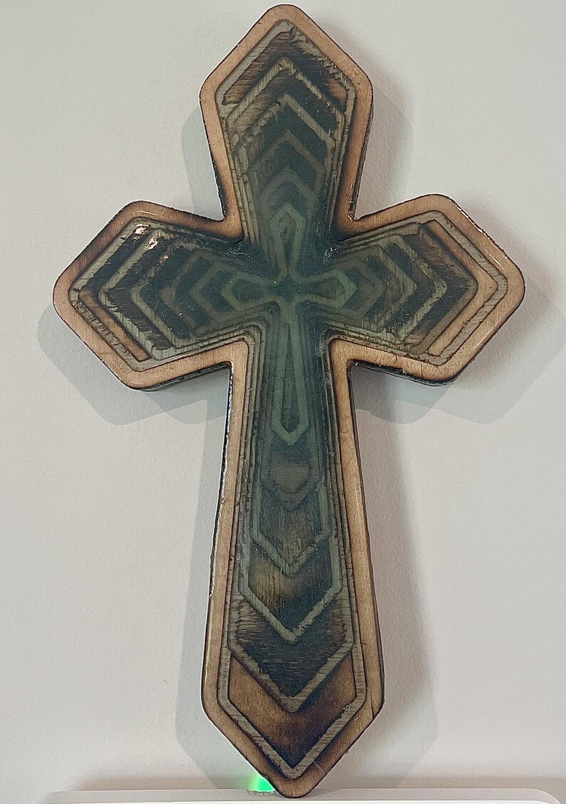 May include: Wooden cross with a layered design. The cross features a light brown border and a central design with shades of green and brown. The cross has a detailed, geometric pattern and is likely a decorative item.