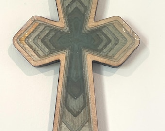 May include: A layered wooden cross with a geometric design. The cross features multiple layers of wood in shades of gray, green, and tan, creating a dimensional effect. The edges are outlined in a darker brown, and the cross is set against a white background.