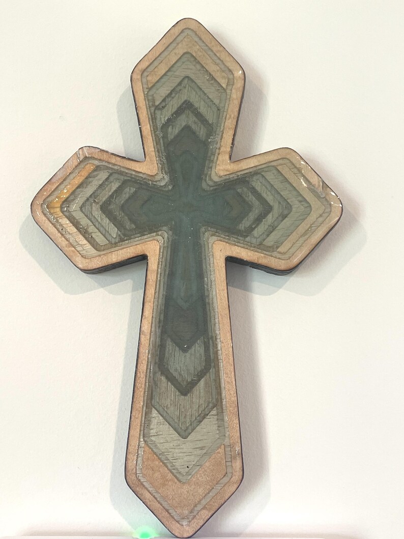 May include: A layered wooden cross with a geometric design. The cross features multiple layers of wood in shades of gray, green, and tan, creating a dimensional effect. The edges are outlined in a darker brown, and the cross is set against a white background.
