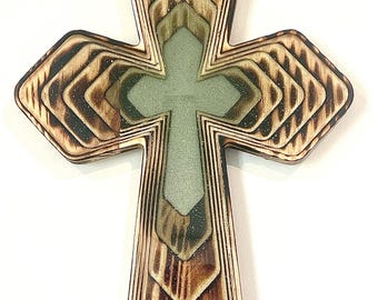 May include: Wooden cross with a layered design. The cross features a light brown wood with darker, burnt edges. The center of the cross is a lighter green color. The cross is a religious symbol.
