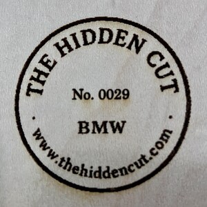 May include: Circular stamp with the text "THE HIDDEN CUT" around the perimeter. The center includes "No. 0029" and "BMW" below it. The website address "www.thehiddencut.com" is at the bottom of the stamp.