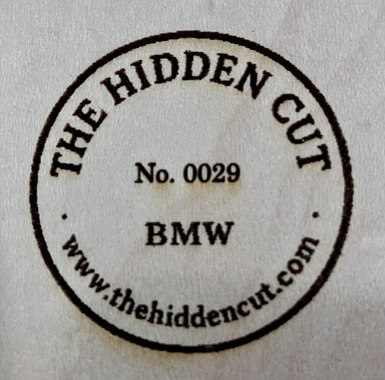 May include: Circular stamp with the text "THE HIDDEN CUT" around the perimeter. The center includes "No. 0029" and "BMW" below it. The website address "www.thehiddencut.com" is at the bottom of the stamp.